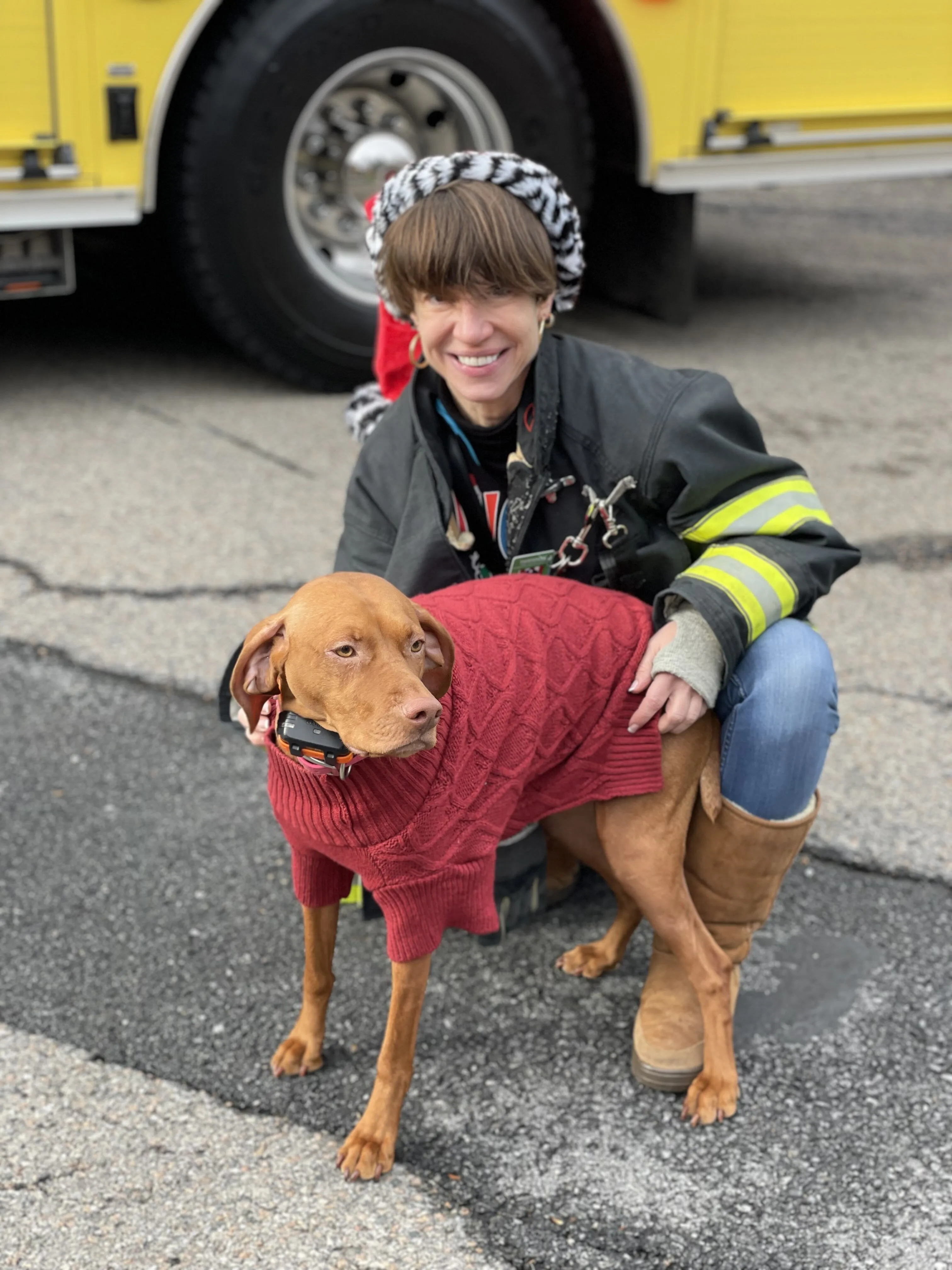 Margaret in turnout gear with a rescue dog in a red sweater in front of a fire engine